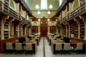 Interno di biblioteca antica con luci accese e tavoli in legno con scaffali e balconate ai lati