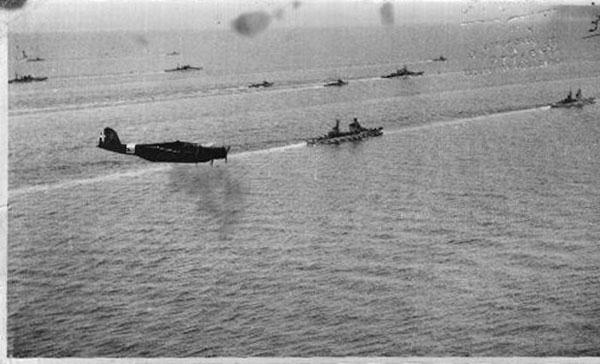 Foto in bianco e nero di un aereo militare che plana e navi da guerra