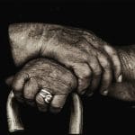 Primo piano delle mani di una donna anziana seduta, appoggiate sul manico ricurvo di un bastone di legno.