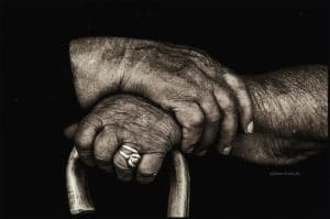 Primo piano delle mani di una donna anziana seduta, appoggiate sul manico ricurvo di un bastone di legno.